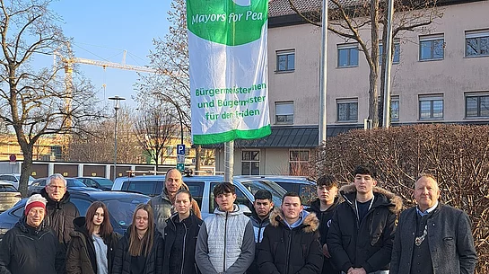 Gruppe von Menschen steht vor einem Fahnenmast mit einer Fahne, die eine Friedenstaube und den Text 'Mayor for Peace' zeigt, im Hintergrund Gebäude und kahler Baum.