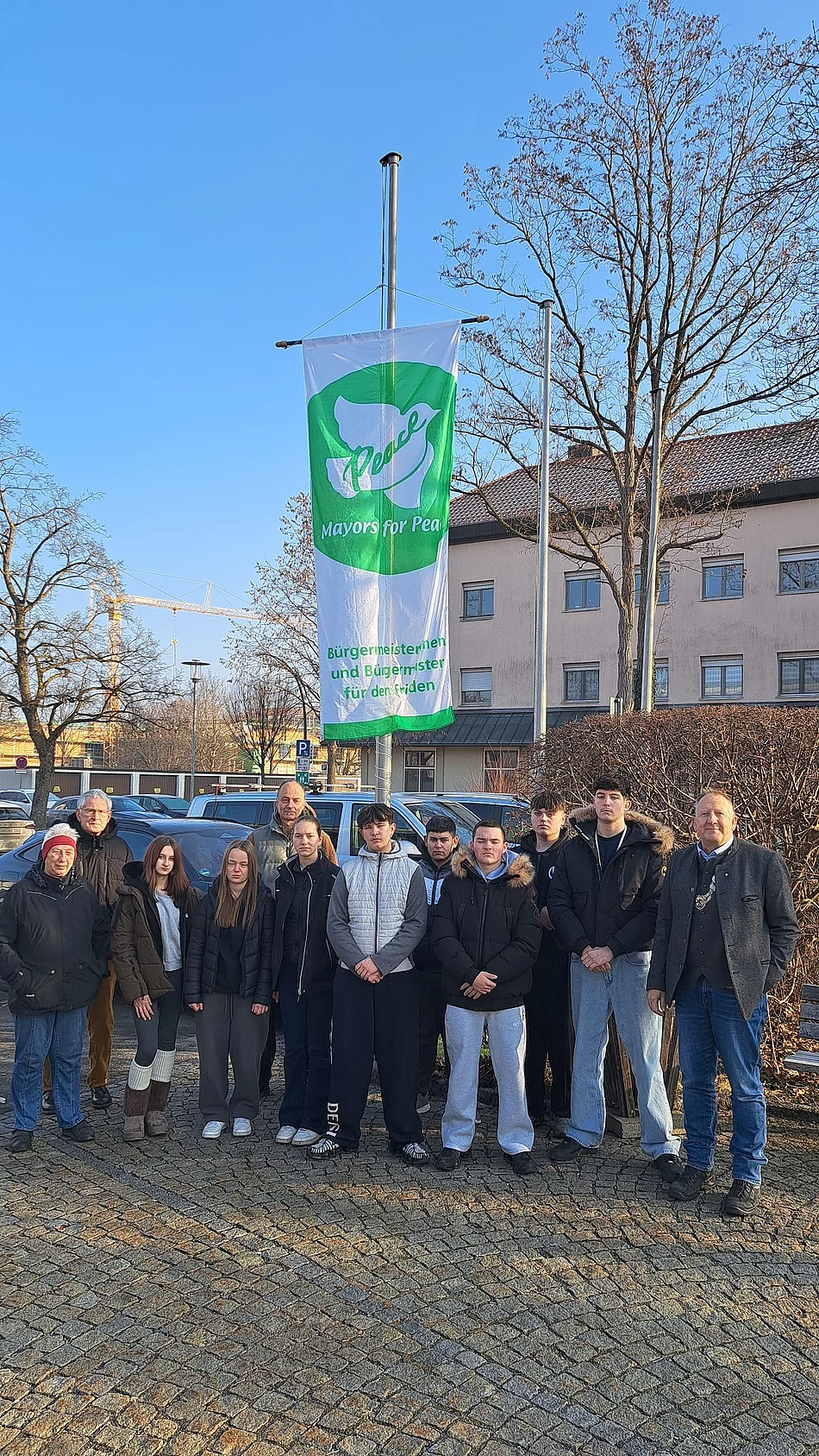 Gruppe von Menschen steht vor einem Fahnenmast mit einer Fahne, die eine Friedenstaube und den Text 'Mayor for Peace' zeigt, im Hintergrund Gebäude und kahler Baum.