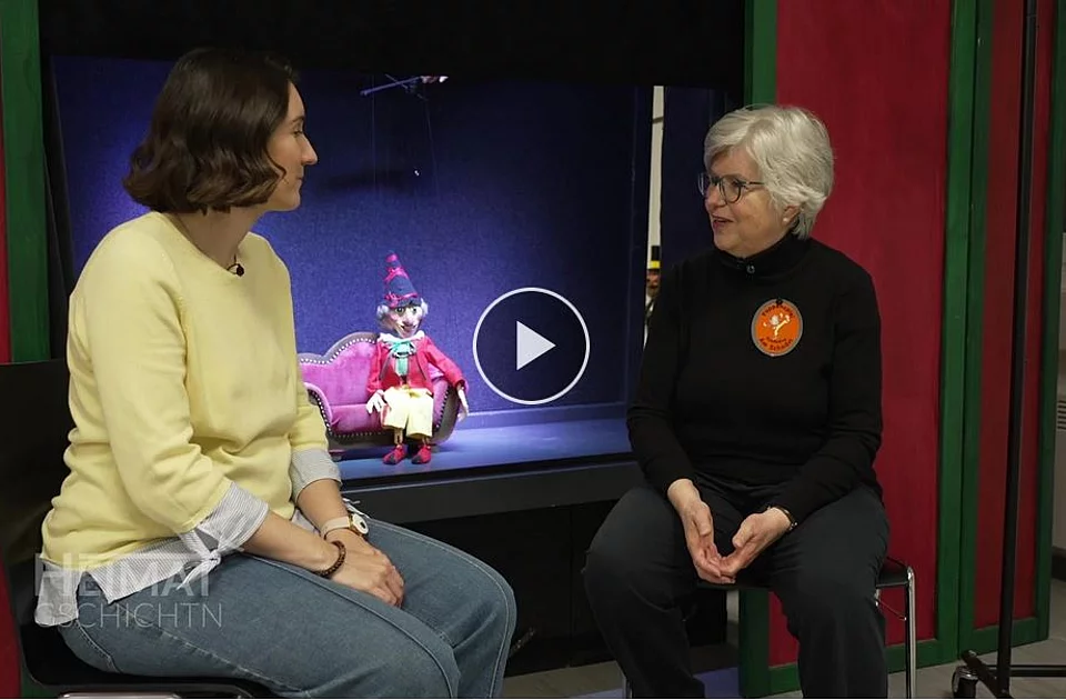 Zwei Frauen sitzen sich in einem Raum mit rotem Vorhang und schwarzem Hintergrund gegenüber, zwischen ihnen steht eine bunte Marionette auf einem kleinen Tisch
