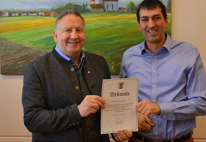 Zwei Männer stehen vor einem Gemälde einer Kirche in einer Landschaft und halten gemeinsam eine Urkunde