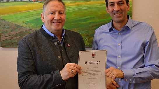Zwei Männer stehen vor einem Gemälde einer Kirche in einer Landschaft und halten gemeinsam eine Urkunde