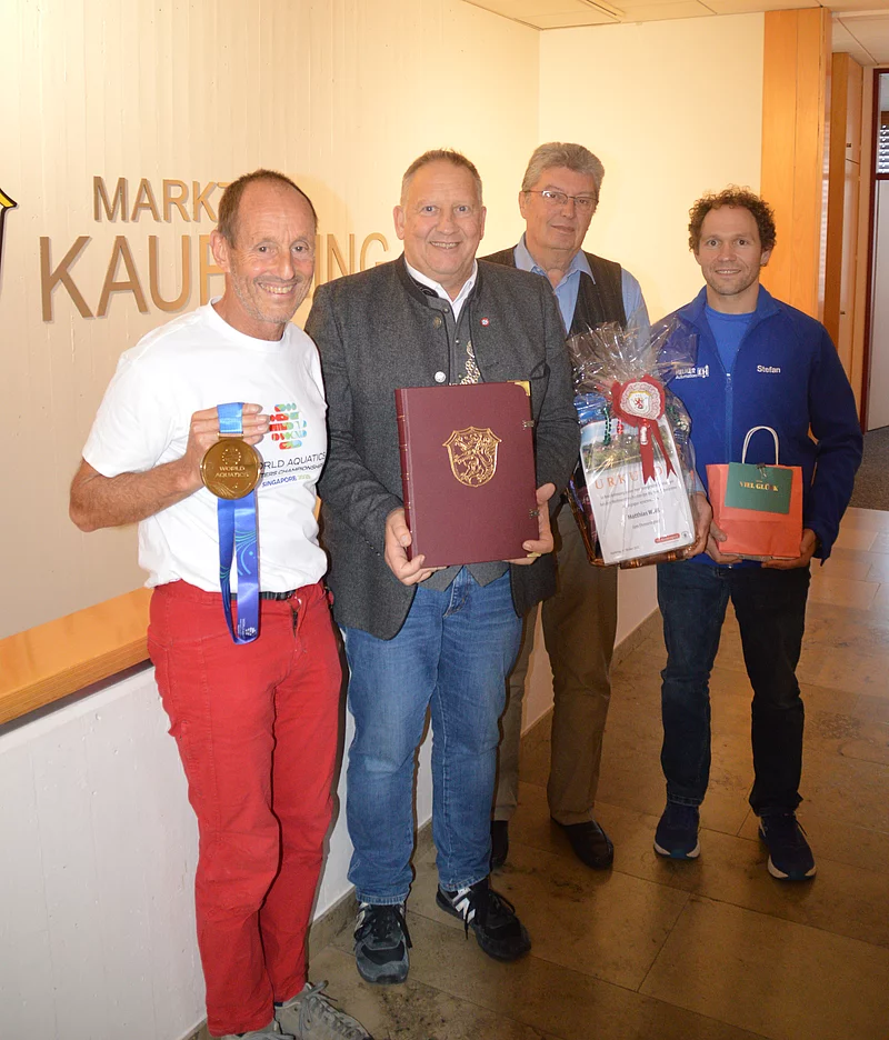 Vier Männer stehen in einem Flur, einer hält eine Medaille mit blauem Band, ein anderer ein rotes Buch mit Wappen, ein weiterer ein Geschenk mit Schleife, der vierte eine kleine Tasche.