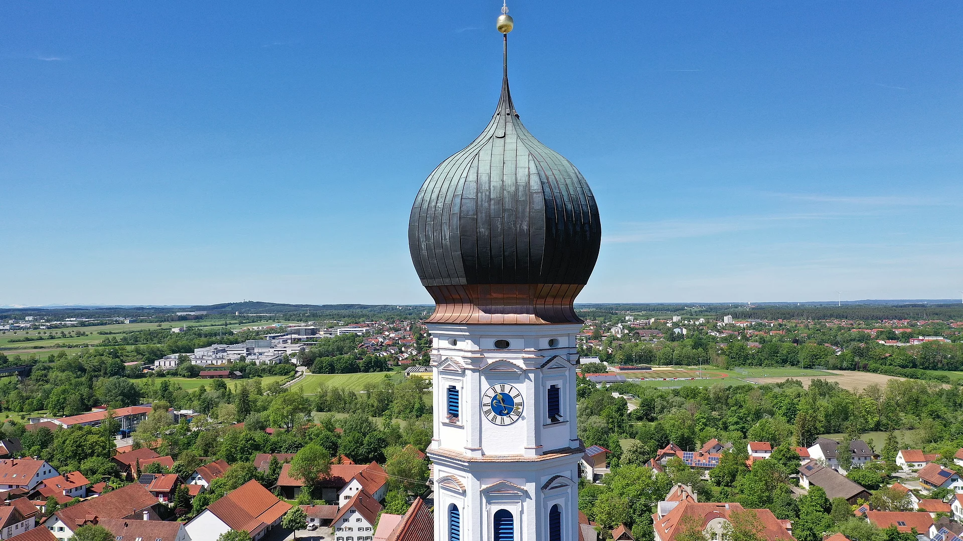 Blick auf den Kirchturm der Pfarrkirche St. Johann mit Uhr-Ziffernblatt. Dahinter sieht man in die Ortschaft Kaufering und sogar bis nach Kaufering Mitte.