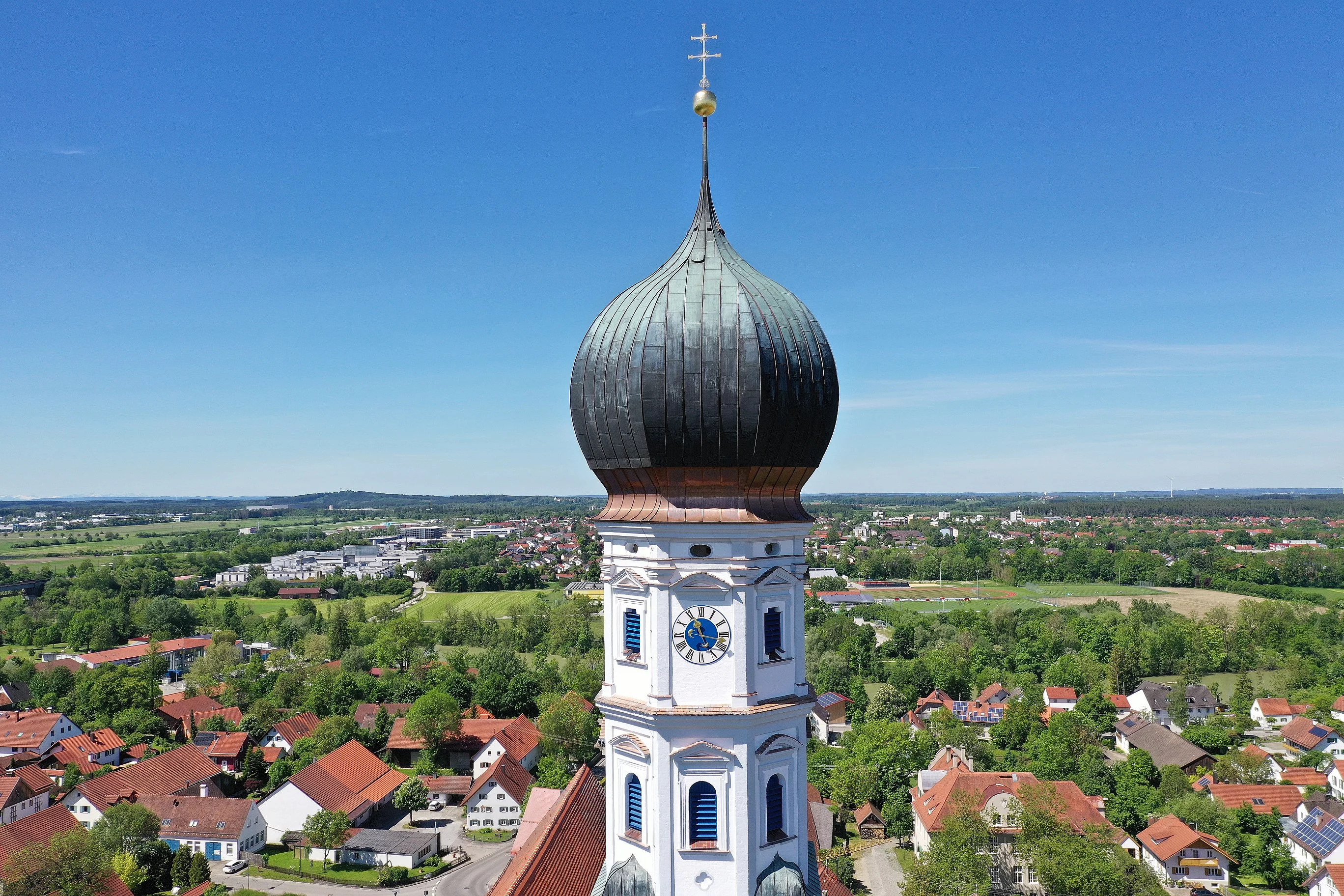 Blick auf den Kirchturm der Pfarrkirche St. Johann mit Uhr-Ziffernblatt. Dahinter sieht man in die Ortschaft Kaufering und sogar bis nach Kaufering Mitte.