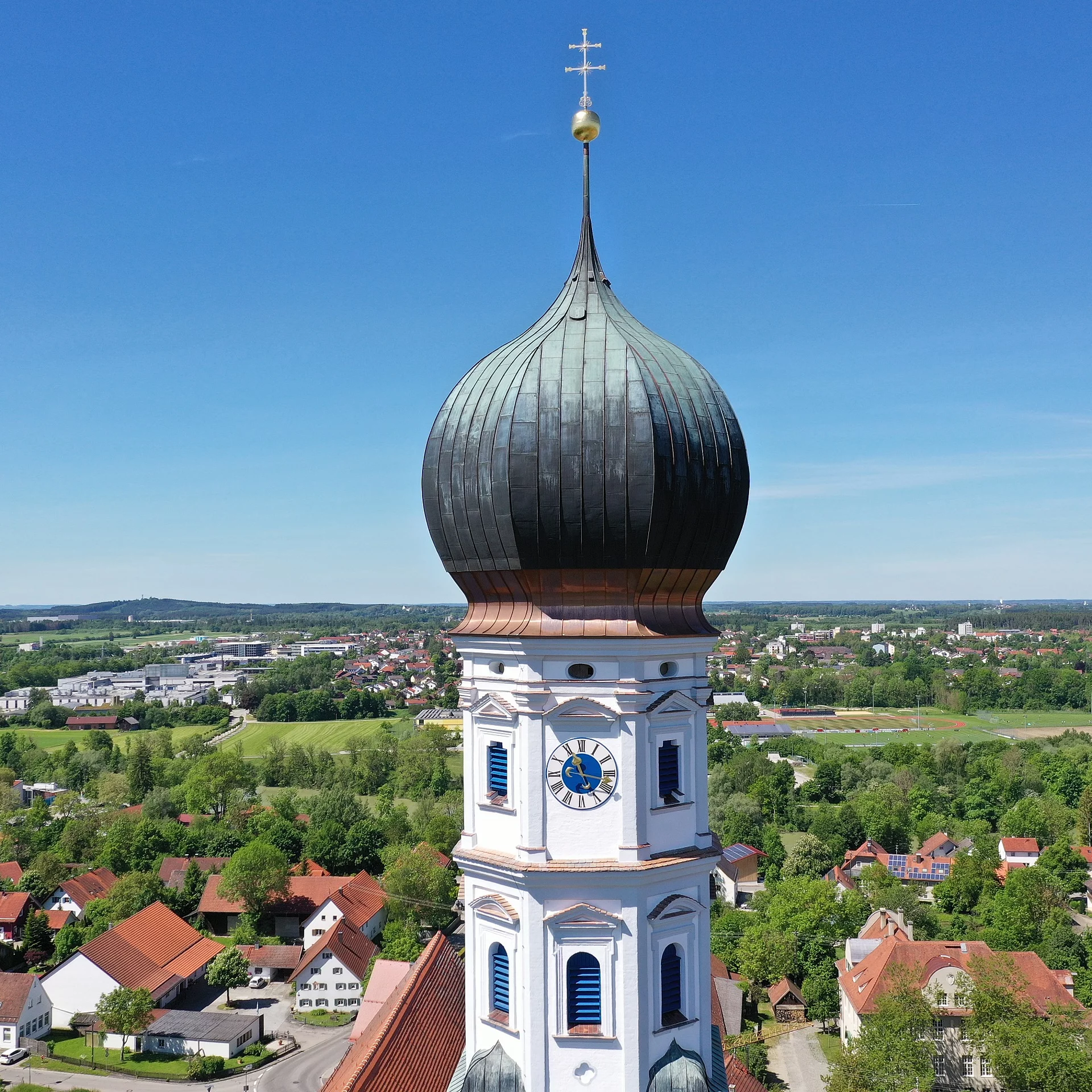Blick auf den Kirchturm der Pfarrkirche St. Johann mit Uhr-Ziffernblatt. Dahinter sieht man in die Ortschaft Kaufering und sogar bis nach Kaufering Mitte.