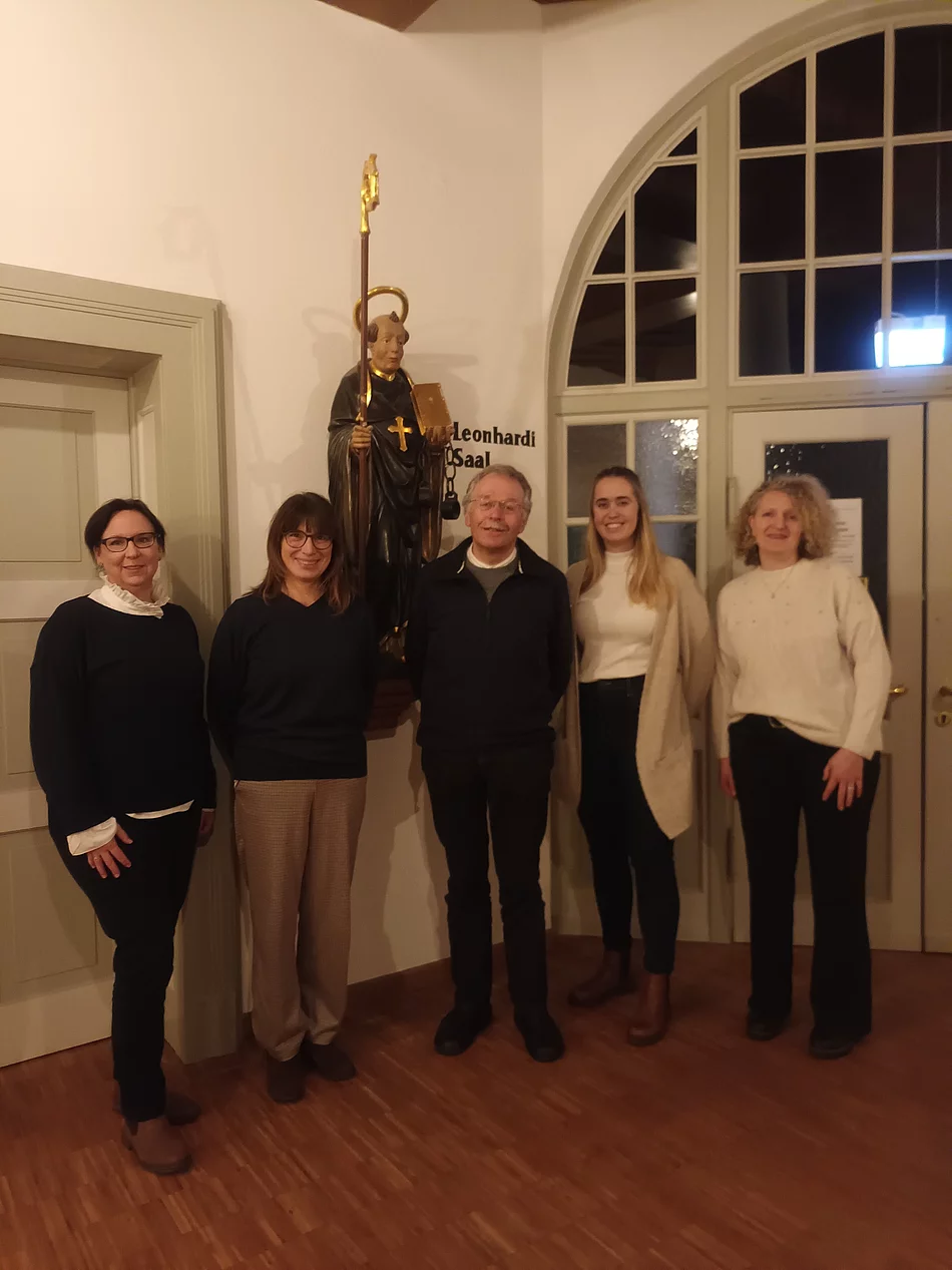 Fünf Personen stehen in einem Raum vor einer Statue mit Bischofsstab und Kreuz, im Hintergrund ein Fenster mit Rundbogen und eine Tür.