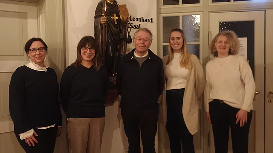 Fünf Personen stehen in einem Raum vor einer Statue mit Bischofsstab und Kreuz, im Hintergrund ein Fenster mit Rundbogen und eine Tür.