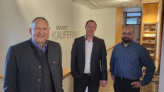 Drei Männer stehen in einem Flur vor einer Wand mit der Aufschrift 'MARKT KAUFERING'.