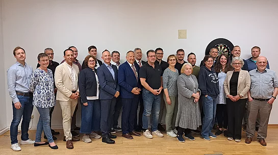 Gruppe von Menschen (neugewählter Marktgemeinderat) in einem Raum vor einer Dartscheibe an der Wand