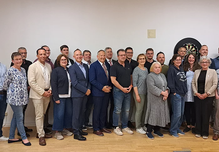 Gruppe von Menschen (neugewählter Marktgemeinderat) in einem Raum vor einer Dartscheibe an der Wand