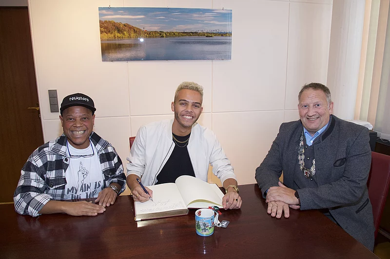 Drei Personen sitzen an einem Holztisch, die mittlere Person schreibt in ein aufgeschlagenes Buch, an der Wand hängt ein Landschaftsfoto mit See und Bäumen.