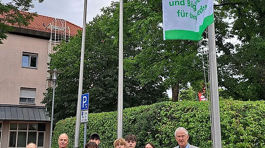 Gruppe von neun Personen steht vor einem Fahnenmast mit einer grünen und weißen Flagge, auf der eine Friedenstaube und der Text 'Mayor for peace'