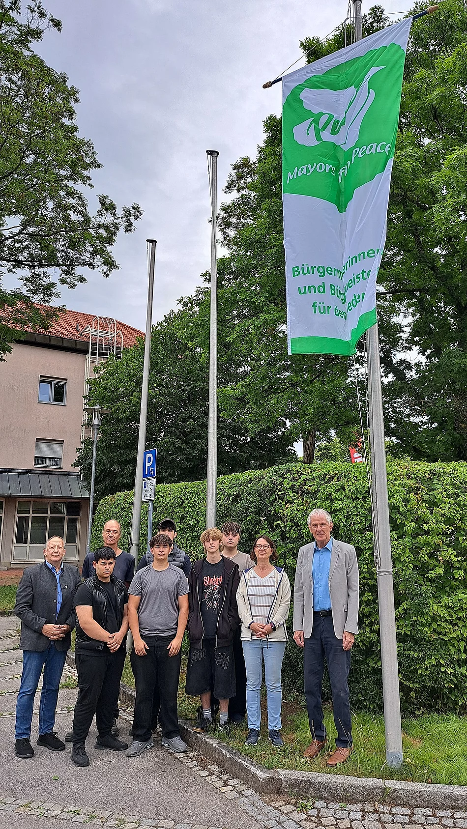 Gruppe von neun Personen steht vor einem Fahnenmast mit einer grünen und weißen Flagge, auf der eine Friedenstaube und der Text 'Mayor for peace' 
