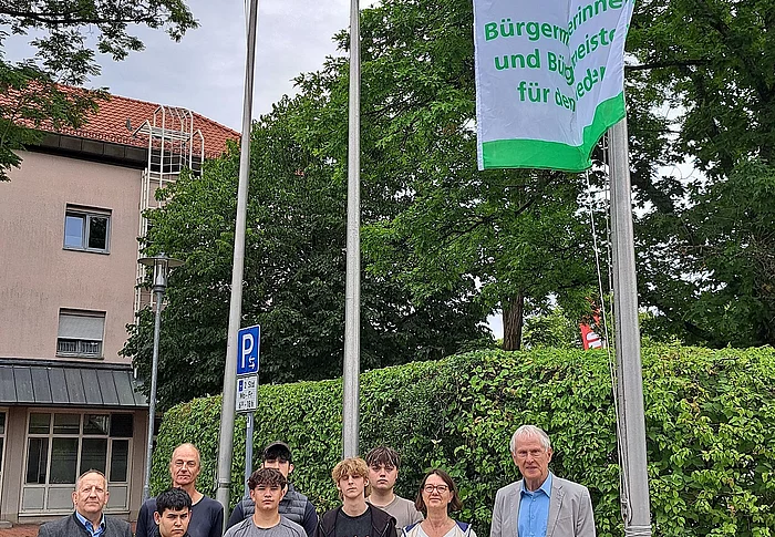 Gruppe von neun Personen steht vor einem Fahnenmast mit einer grünen und weißen Flagge, auf der eine Friedenstaube und der Text 'Mayor for peace' 