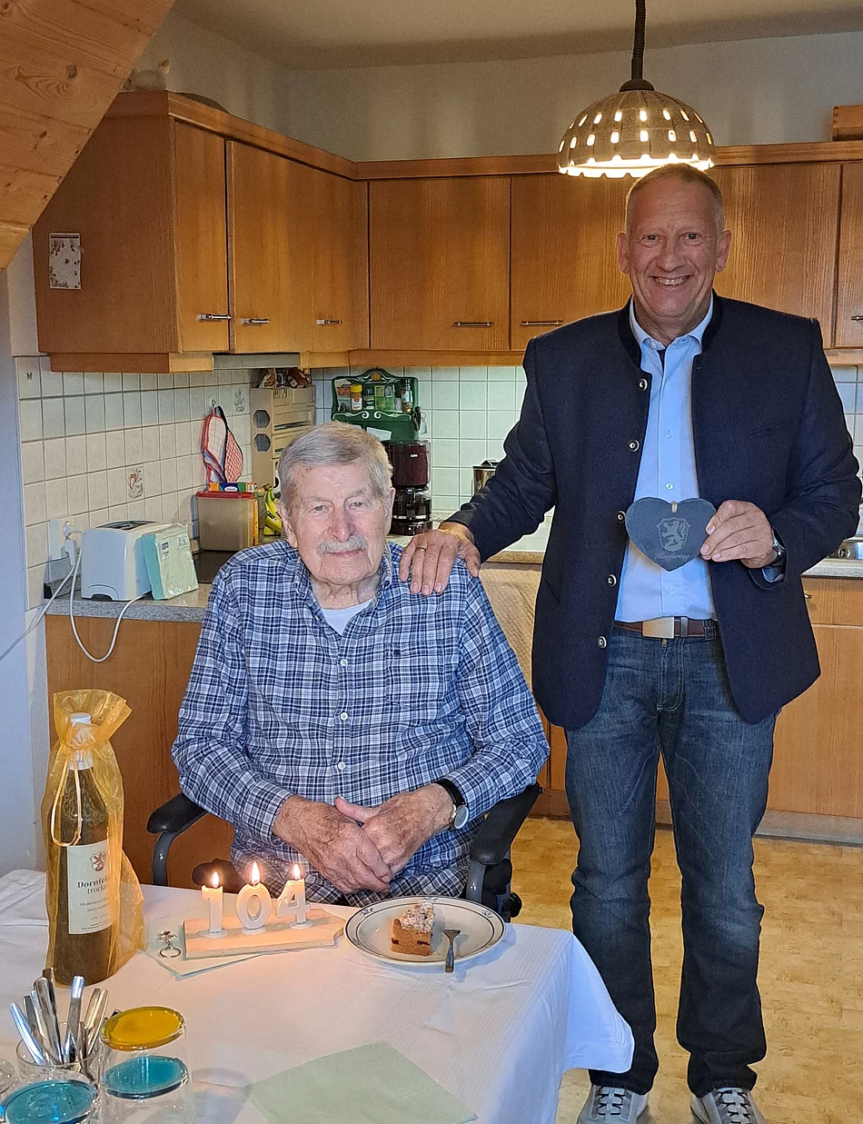 Zwei Männer in einer Küche, einer sitzt am Tisch mit Geburtstagskerzen und Kuchen, der andere steht daneben und hält ein Herz aus Stoff.
