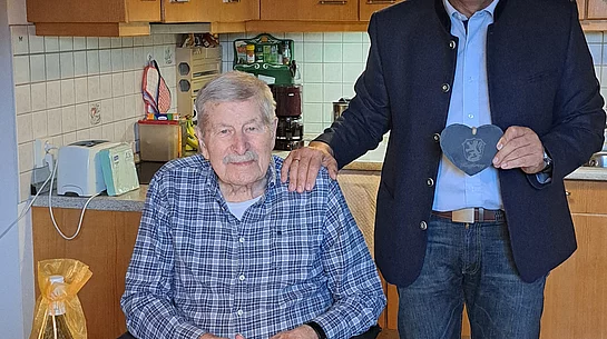 Zwei Männer in einer Küche, einer sitzt am Tisch mit Geburtstagskerzen und Kuchen, der andere steht daneben und hält ein Herz aus Stoff.