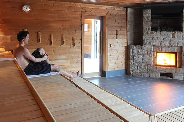 Lechtalbad Kaufering - Kaminsauna - Frau sitzt mit schwarzem Handtuch bedeckt auf der Sitzbank vor dem Kaminfenster in der Sauna