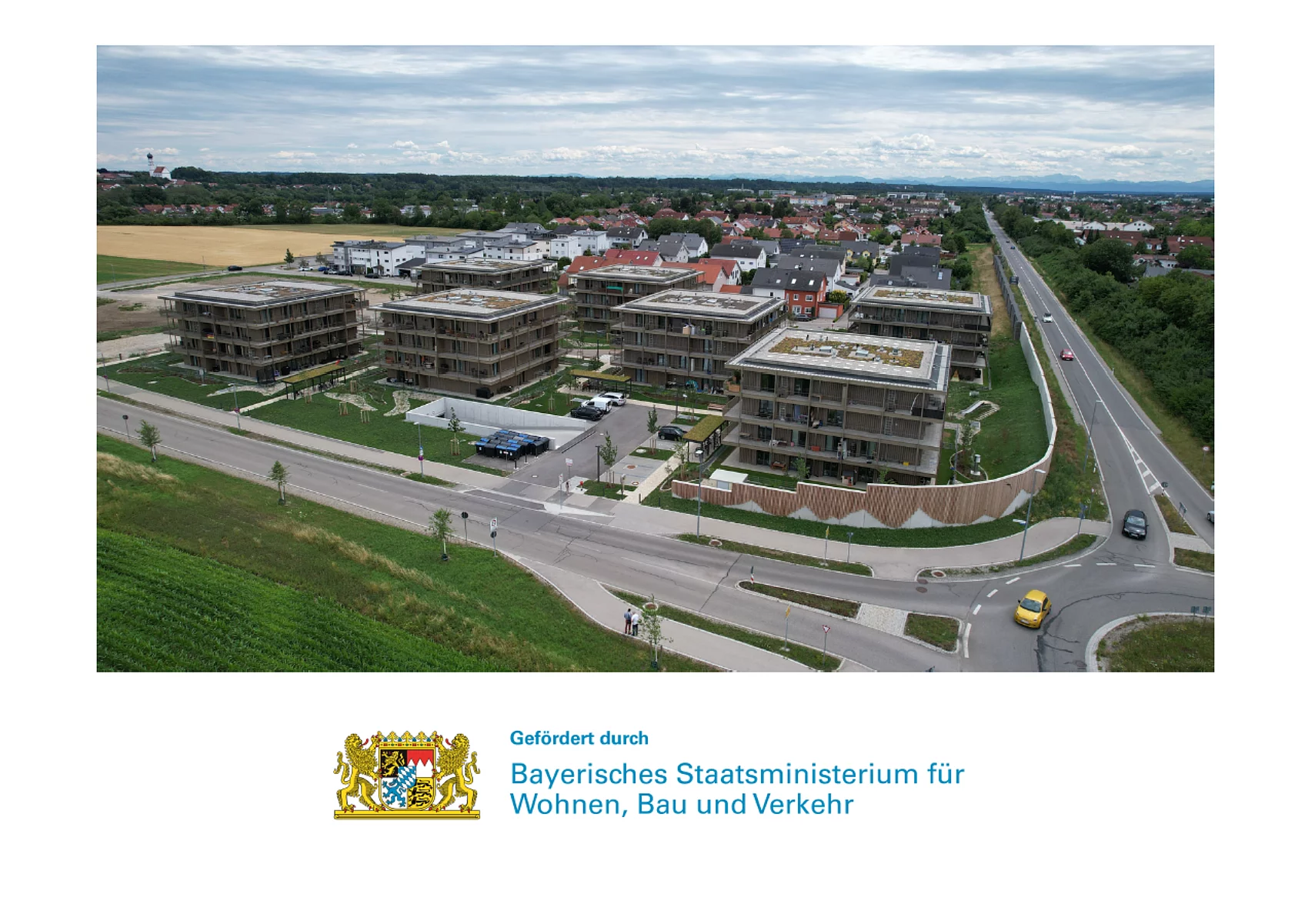Luftaufnahme von mehreren modernen Wohngebäuden an einer Kreuzung mit angrenzenden Feldern und einer Straße mit Fahrzeugen