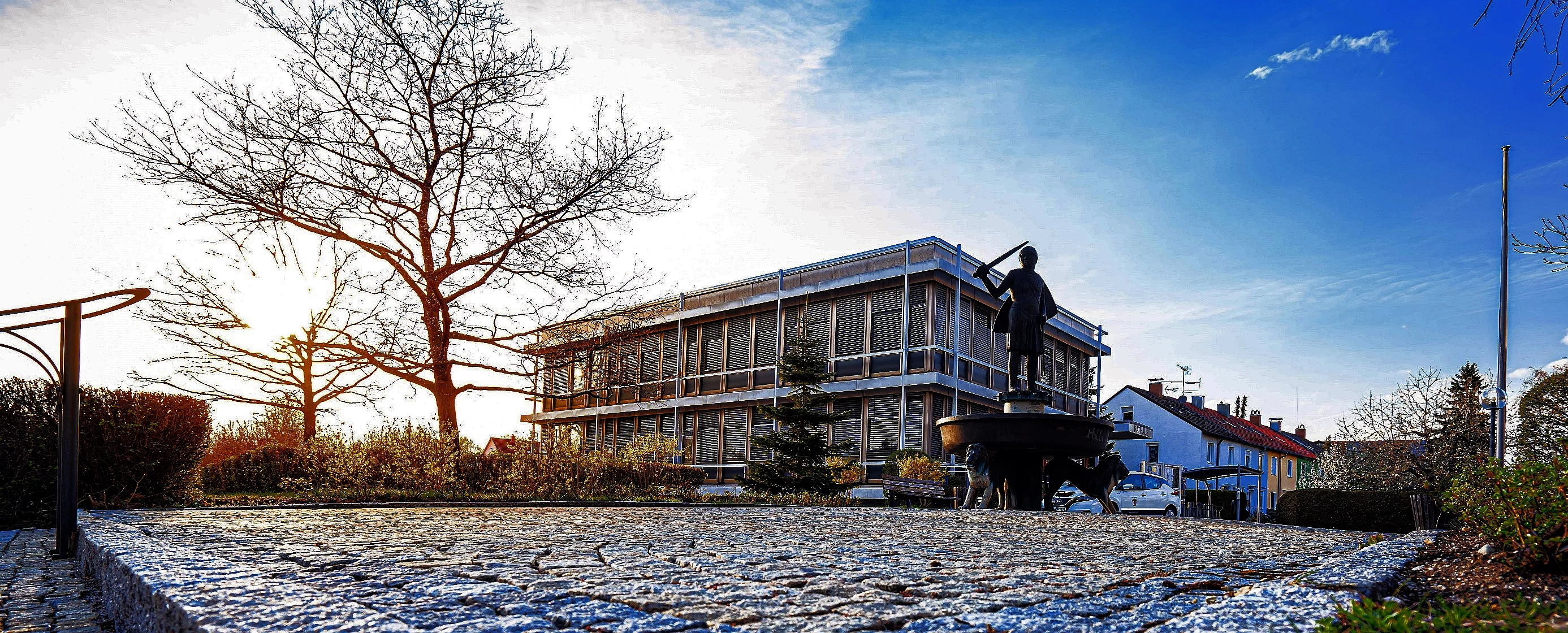 Blick auf das Rathaus, das in der Frühlingssonne erhellt wird. vorne ein gepflasterter Platz mit dem Welfenbrunnen. Im Hintergrund das Rathaus. Abends strahlt die Sonne durch den einzelnen blattlosen Baum.