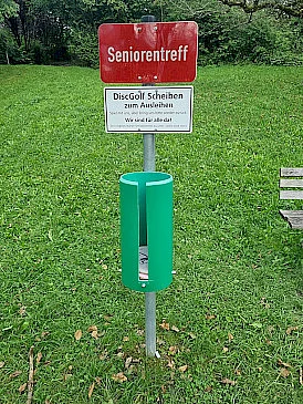 DiscGolf Ausleihstation - Rotes Schild mit Seniorentreff-Schild und grünem Behälter mit den Discscheiben