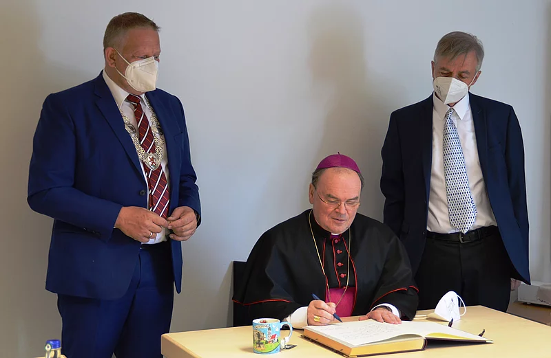 Ein Mann in Bischofsgewand sitzt an einem Tisch und schreibt in ein großes Buch, zwei Männer in Anzügen stehen daneben.
