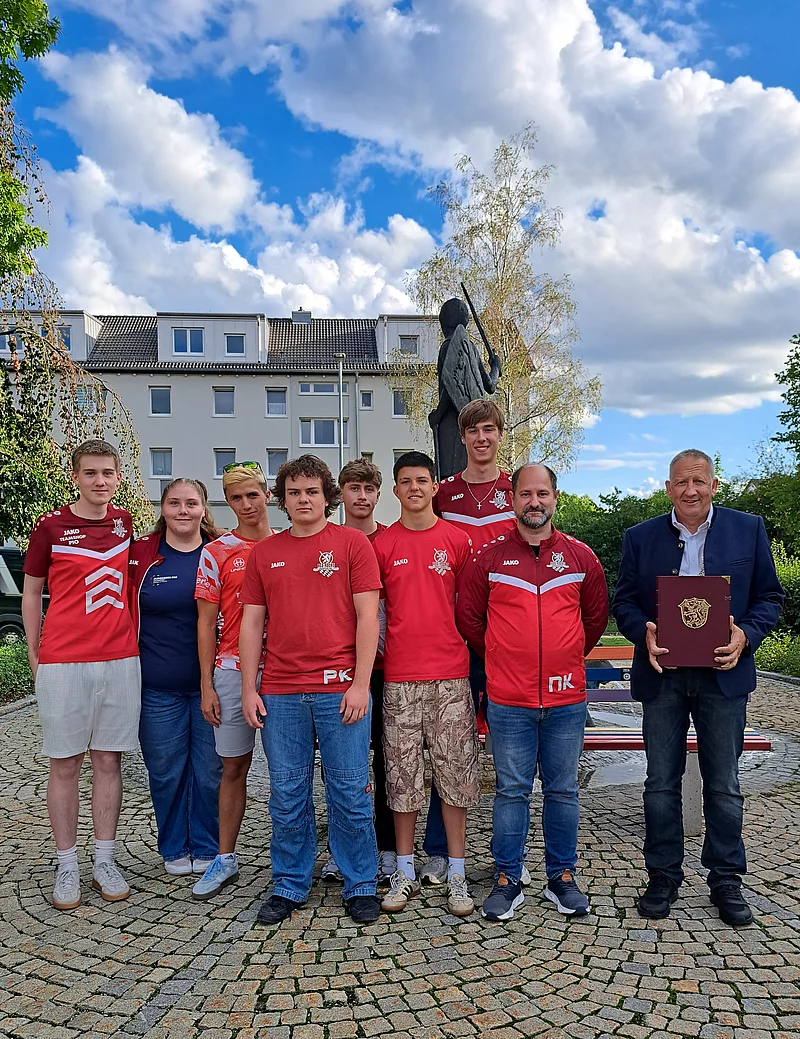 Gruppe von neun Personen steht vor einer Statue auf einem gepflasterten Platz unter blauem Himmel mit Wolken. Die Person rechts hält ein Buch in den Händen.