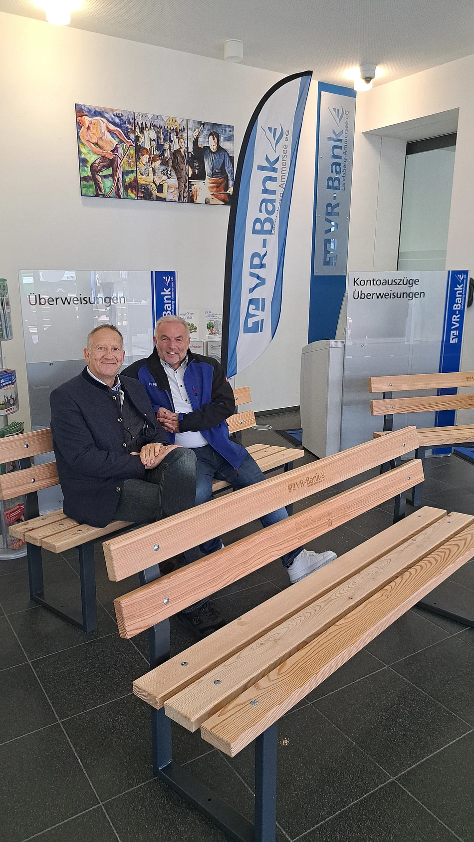 Zwei Männer sitzen auf einer Holzbank in einem Raum mit VR-Bank-Werbefahnen und Informationsständern zu Kontoauszügen und Überweisungen