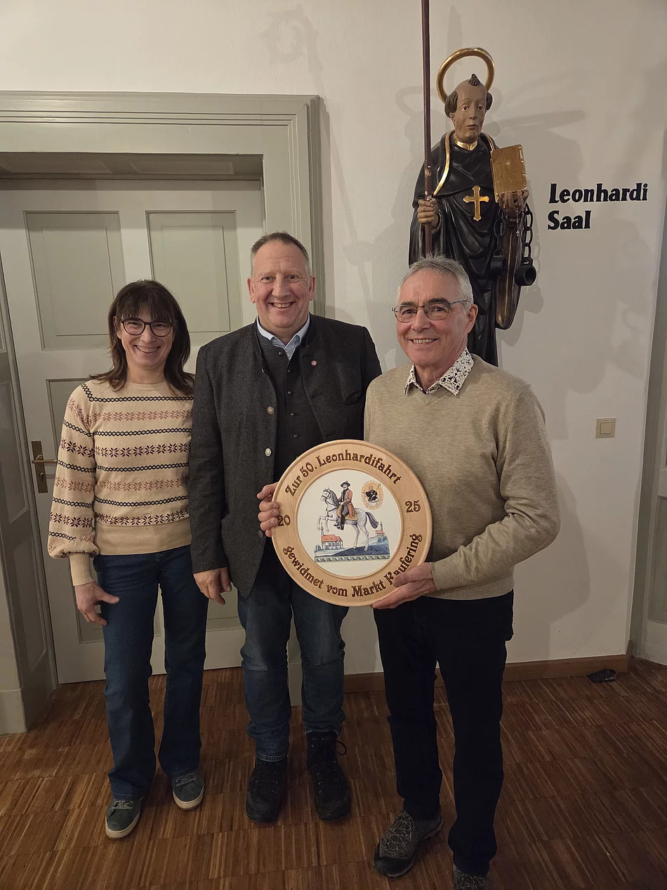Drei Personen stehen vor einer Statue eines Heiligen mit Stab und Buch im Raum mit der Aufschrift 'Leonhardi Saal', eine Person hält eine runde Jubiläumsholzscheibe mit bemaltem Motiv und Schriftzug