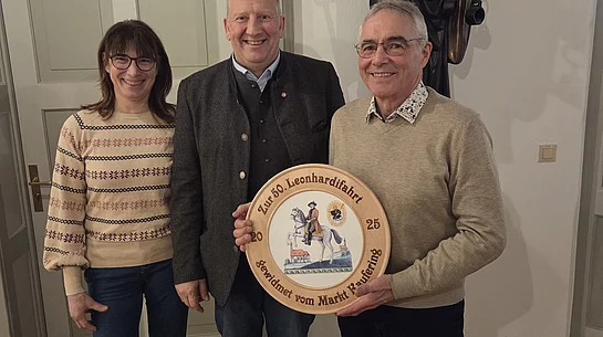 Drei Personen stehen vor einer Statue eines Heiligen mit Stab und Buch im Raum mit der Aufschrift 'Leonhardi Saal', eine Person hält eine runde Holzscheibe mit bemaltem Motiv und Schriftzug