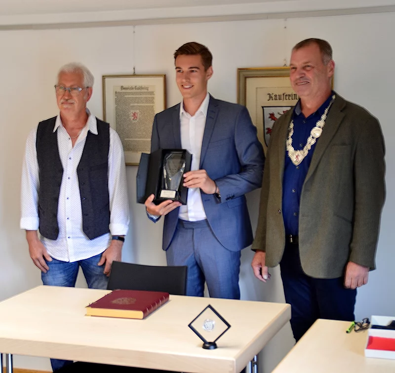 Drei Männer stehen hinter einem Tisch, einer hält eine Auszeichnung in einer Box, auf dem Tisch liegen ein großes Buch und ein weiterer Preis in einem Ständer.