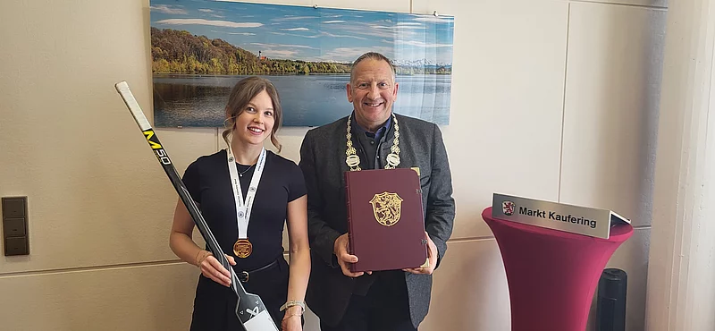 Zwei Personen stehen nebeneinander, die linke hält einen Hockeyschläger und trägt eine Medaille, die rechte hält eine Urkunde mit Wappen, im Hintergrund ein Bild eines Sees und rechts ein Stehtisch mit Schild Markt Kaufering