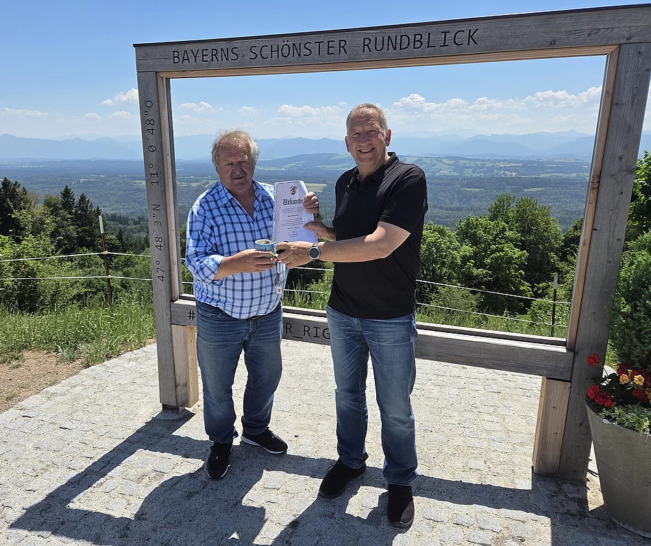 Zwei Männer stehen vor einem großen Holzrahmen mit der Aufschrift 'BAYERNS SCHÖNSTER RUNDBLICK', einer hält eine Urkunde, die andere eine Tasse, im Hintergrund Landschaft mit Bergen und blauem Himmel