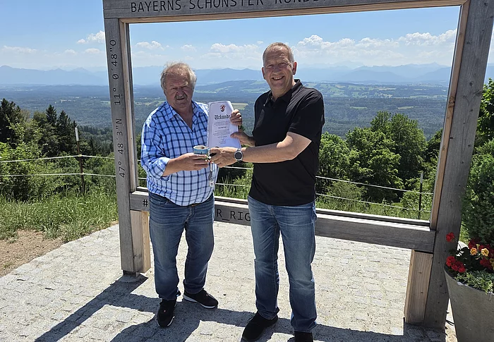 Zwei Männer stehen vor einem großen Holzrahmen mit der Aufschrift 'BAYERNS SCHÖNSTER RUNDBLICK', einer hält eine Urkunde, die andere eine Tasse, im Hintergrund Landschaft mit Bergen und blauem Himmel