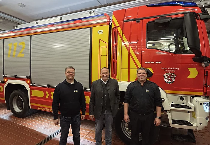 Drei Männer stehen vor einem roten und gelben Feuerwehrfahrzeug mit der Nummer 112 in einer Fahrzeughalle