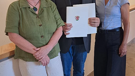 Drei Personen stehen nebeneinander in einem Raum mit heller Wand und Wappen, die mittlere Person hält ein Dokument mit einem Wappen