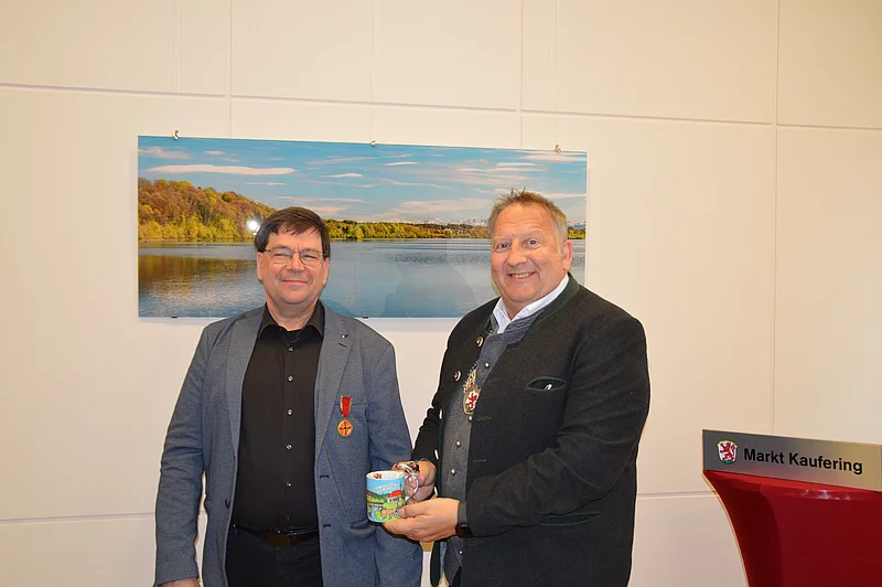 Zwei Männer stehen nebeneinander, einer trägt eine Medaille, der andere hält eine Tasse. Im Hintergrund hängt ein großes Landschaftsfoto mit See und Bäumen.