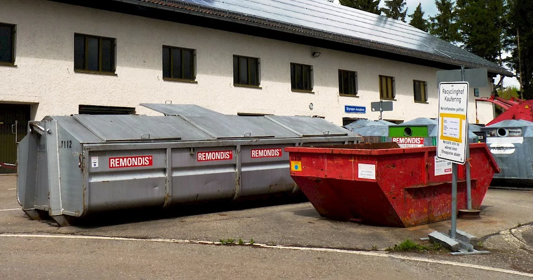 Verschiedene rote und silberne Abfall Container stehen vor dem Gebäude des Recyclinghofs