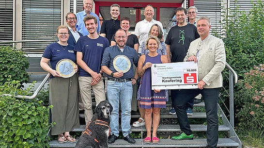 Gruppe von Menschen steht auf einer Treppe vor einem Gebäude, zwei Personen halten große Münznachbildungen, eine Person hält einen großen Scheck, ein Hund sitzt vorne auf der Treppe